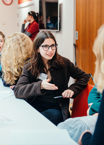 A smiling young woman is sat at a table, turned towards someone else who is off camera. They are in conversation. Other people can be seen in the background, also sat at the table talking.
