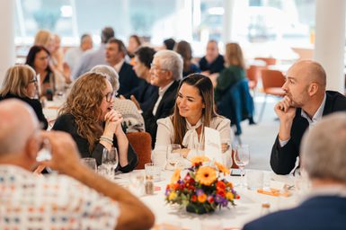 A group of people sit together around a table at a business lunch. They wear smart clothes and a lady and a man listen intently to another lady who is speaking enthusiastically. The table is neatly laid with a white cloth, colourful floral centrepiece, cutlery and glasses.