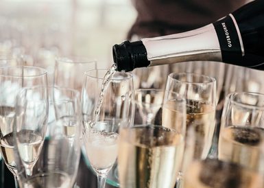 A close up of rows of glasses of sparkling wine. A bottle is being poured, with a label that reads 'Gusbourne'.