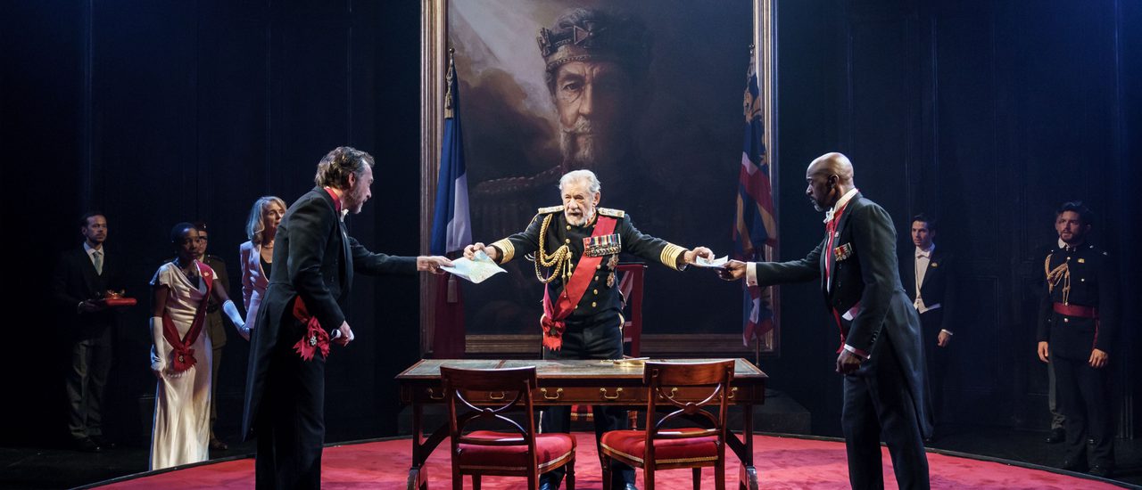 King Lear stands centre, wearing military blues decorated with medals, behind a wooden table and chairs, holding out papers to two men in formal dress with their backs to the photographer. There is a large painting of the king looming over them on the wall, rich red carpets, and others in formal dress looking on from the sides.