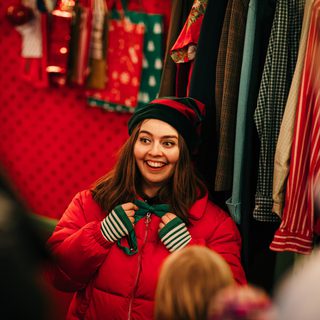 An elf (wearing a red puffer jacket and green and red pointy hat) speaks with a group of visitors to the Grotto.