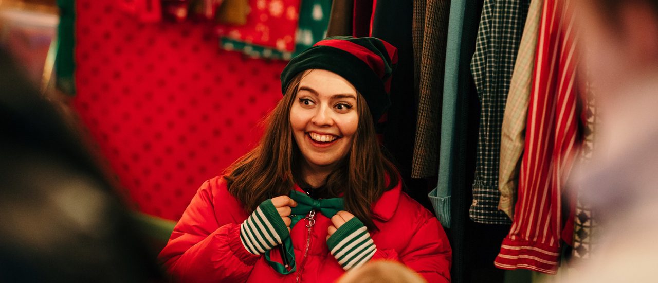 An elf (wearing a red puffer jacket and green and red pointy hat) tweaks her bow tie and looks off to the left. Her face is lit up in the magic of Christmas.