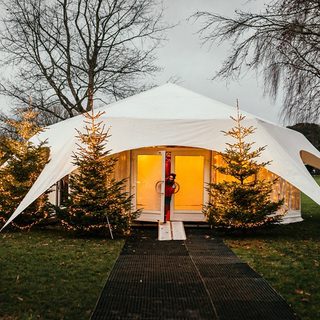 A wintery day with two trees standing tall on the park. In between them the grotto stands, it is a gazebo with white lining over the top reaching to the ground at four visible points nailed into the ground. There are four lit Christmas trees outside the golden illumined doors, an elf stands halfway out of the double doors on the left, smiling at the camera.