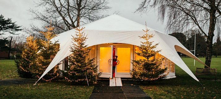 A wintery day with two trees standing tall on the park. In between them the grotto stands, it is a gazebo with white lining over the top reaching to the ground at four visible points nailed into the ground. There are four lit Christmas trees outside the golden illumined doors, an elf stands halfway out of the double doors on the left, smiling at the camera.