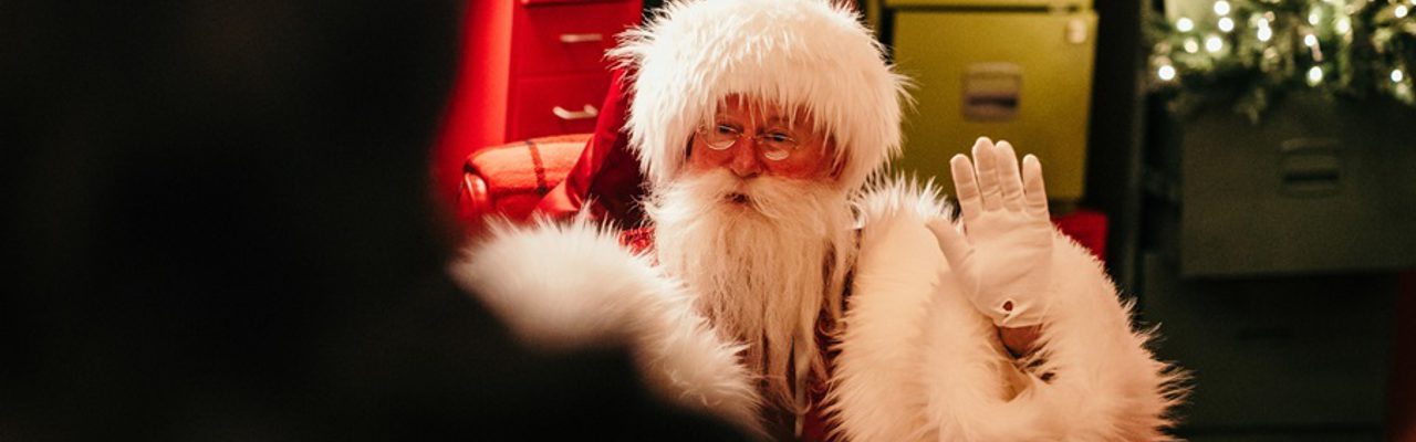 Santa waves from his red arm chair. He wears small spectacles, white gloves, a large red coat with fluffy white trims and has a long fuzzy white beard. Behind him is an assortmenyt of colourful drawer units, one drawer is open and a christmas tree rises out of it with warm lights on its branches.