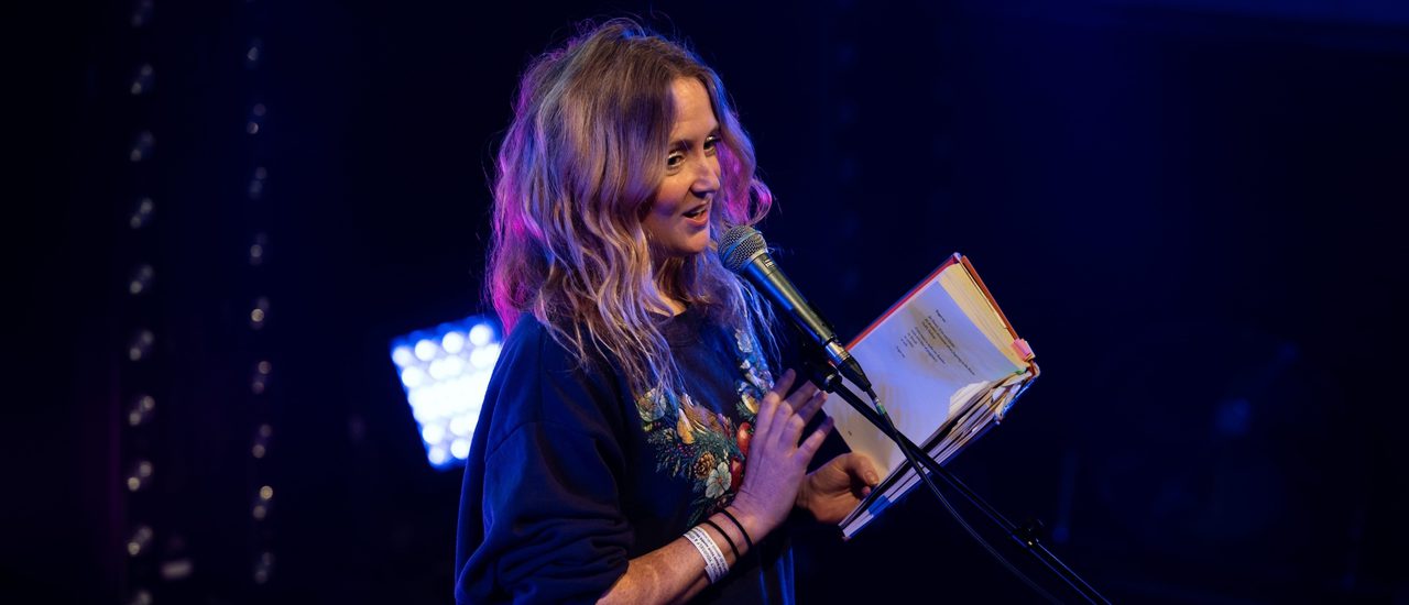 A performer stands in front of a microphone with an open book in their hand. They have long blonde hair and wear a grey graphic t-shirt. They perform the reading with a relaxed smile.