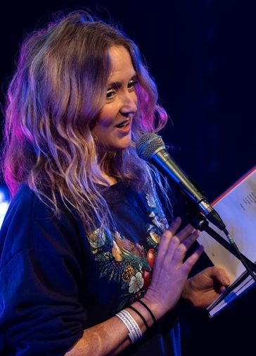 A performer stands in front of a microphone with an open book in their hand. They have long blonde hair and wear a grey graphic t-shirt. They perform the reading with a relaxed smile.