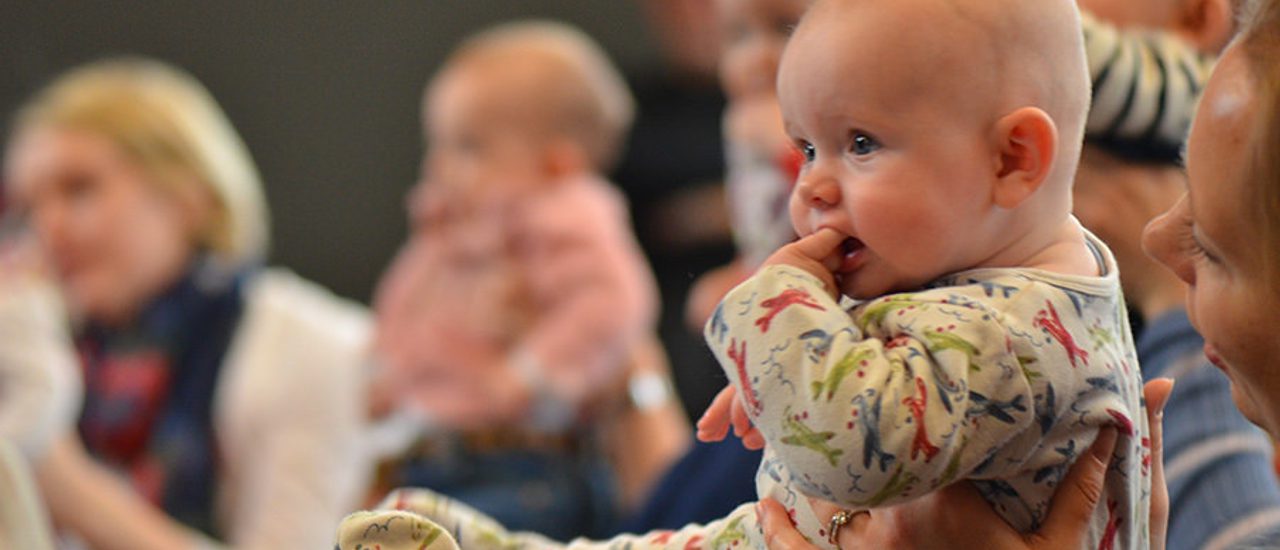 A baby is held up by their parent, with other babies in the background.
