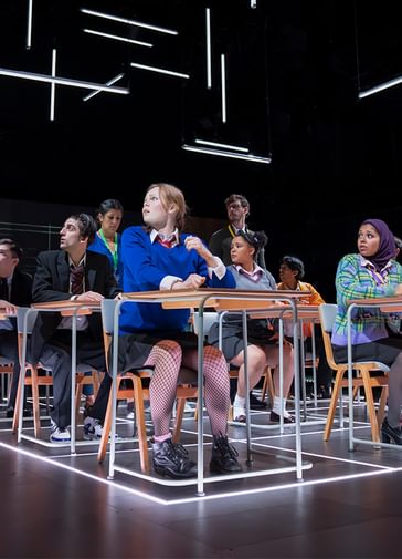 A school classroom with three rows of desks stretching back. Pupils are sitting at the desks, all looking to their right past a teacher on their right, who also looks in the same direction. There is a large clock projected on the back wall and strip lights arranged at various angles above their heads.