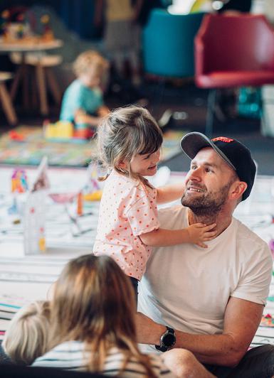 a man is sitting in the theatre foyer on the floor with a toddler. There are other children in the foreground and behind the man there are more children and a floorcloth with paper models.