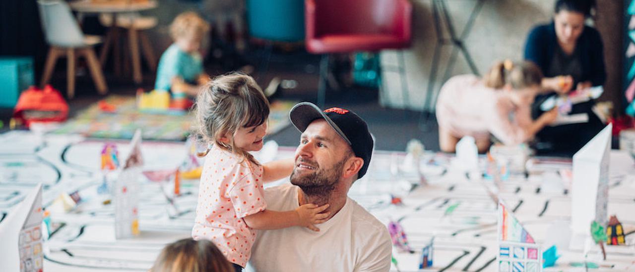 a man is sitting in the theatre foyer on the floor with a toddler. There are other children in the foreground and behind the man there are more children and a floorcloth with paper models.
