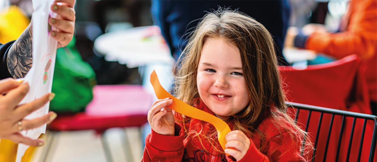 A girl in a red jumper smiles at the camera. She is holding orange tissue paper. Two hands are seen holding a piece of white paper with drawings on it in the left of the frame.