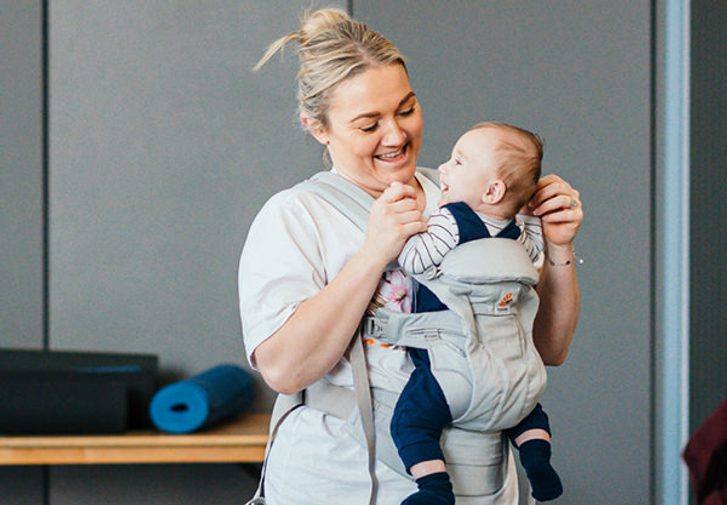 A woman and a little baby in a sling dance while looking and smiling at each other.