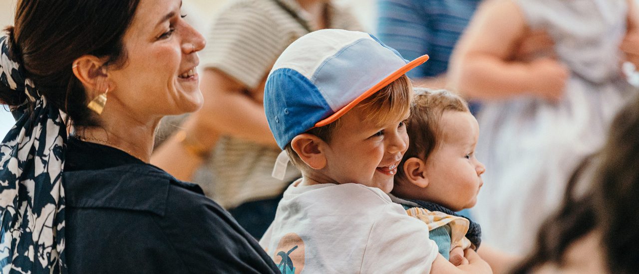 A young boy in a blue baseball cap smileswhile holding a baby. Next to the boy a woman in a black shirt, with a gold earing and white hair tie smiles.