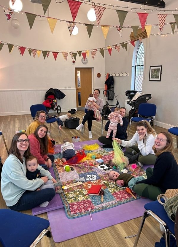 A group of women and babies and toddles sat in a circle smiling warmly at the camera.