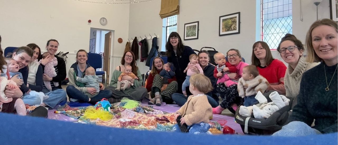 A groups of women and babies and toddles sat in a circle smiling warmly at the camera.