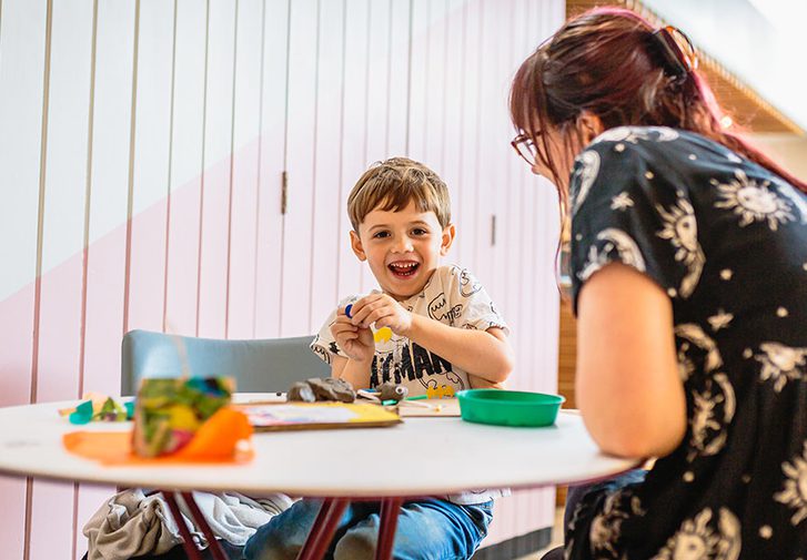 A child and their parent are sat playing with crafts at a table and smiling at the camera.
