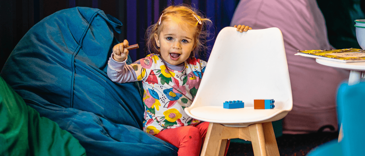 A toddler sits on a beanbag in the foyer of the Theatre. They are holding a crayon.