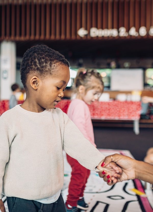 A young boy holds his mother's hand; children play in the background.