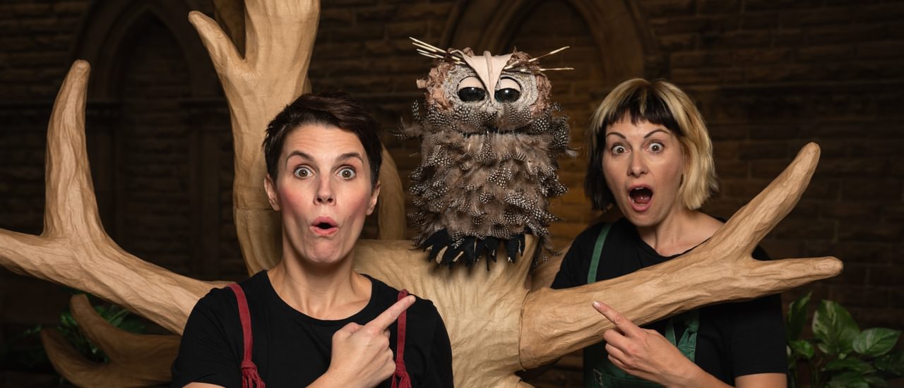 An owl puppet sitting in a tree, two women point towards it looking shocked. They are both wearing black t-shirts and colourful dungarees.