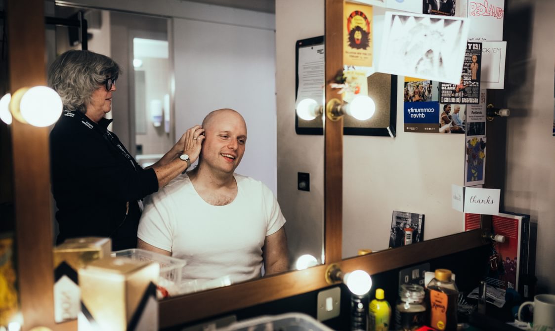 An actor looks in the mirror, smiling as a microphone is fixed to his ear. The man wears a white t-shirt and sits in a chair. Above him, a woman wearing black is touching his ear. Around the mirror are dressing room light bulbs. Many cards have been stuck to the mirror.