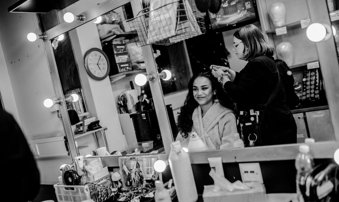 An actor gets ready in the dressing room chair while someone does their hair. Surrounded by various dressing room items, they smile into the mirror. The actor wears a dressing gown and the wig technician wears dark clothes with a tool kit on their hip.