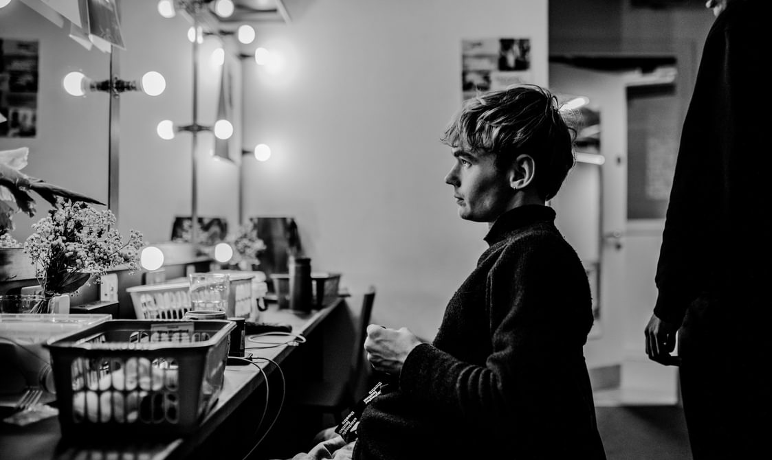 An actor sits in a chair and looks into the dressing room mirror with a neutral expression. The room is lined with mirrors, surrounded by dressing room light bulbs. He wears a relaxed jumper. Behind him, another man is just in shot and looks as if he is talking to the seated actor.