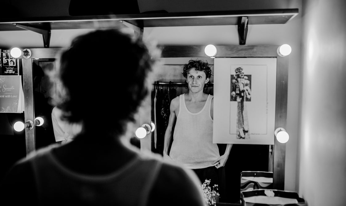An actor stands in the dressing room, looking at himself in the mirror. He has a serious, contemplative expression. The mirror is surrounded by dressing room light bulbs. There is a stylish design sketch picture stuck to the mirror. The actor wears a white tank top.