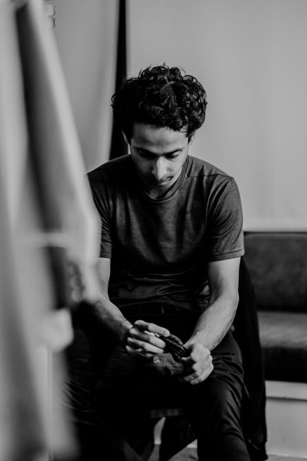 An actor sits alone, looking reflective as he takes a quiet moment. He holds a piece of material in his hands as he looks down at it. He is wearing a dark t-shirt and trousers.