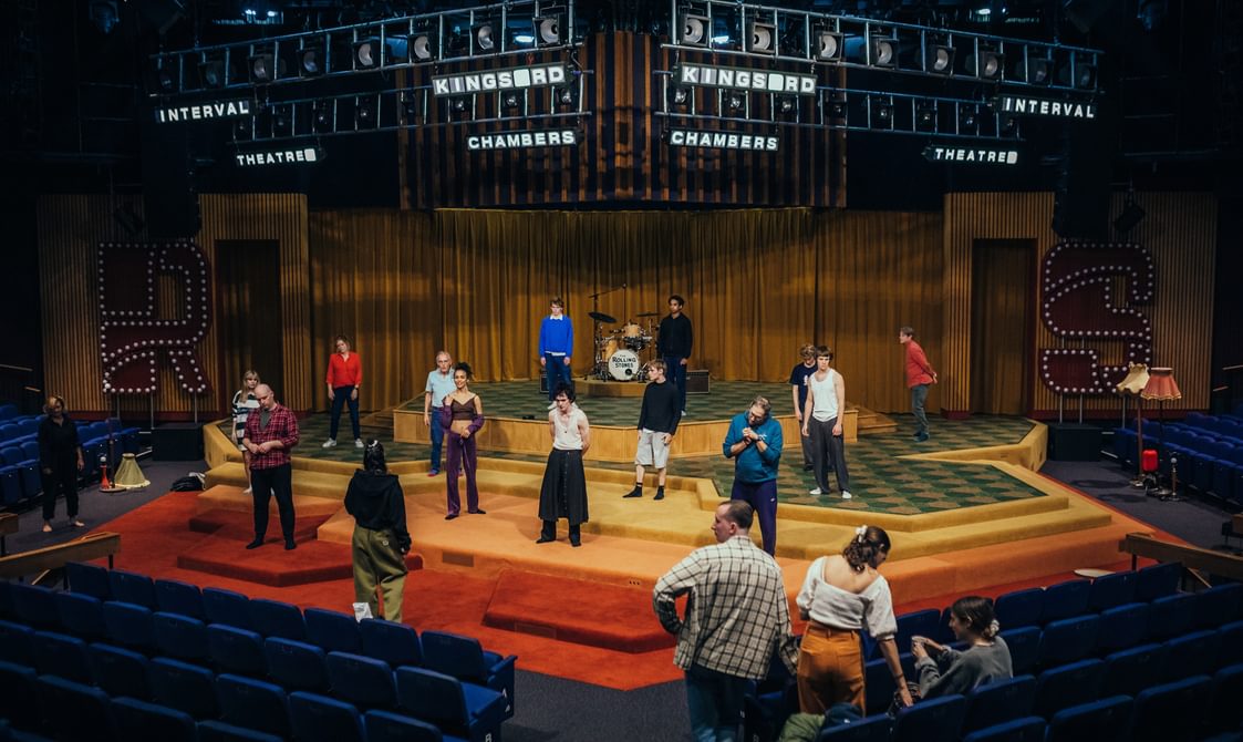 Actors stand on the stage facing towards a woman in the auditorium. The stage is arranged in many hexagonal layers, carpeted in red, orange and yellow. The actors are wearing relaxed clothing. There are a few other people scattered around the auditorium.