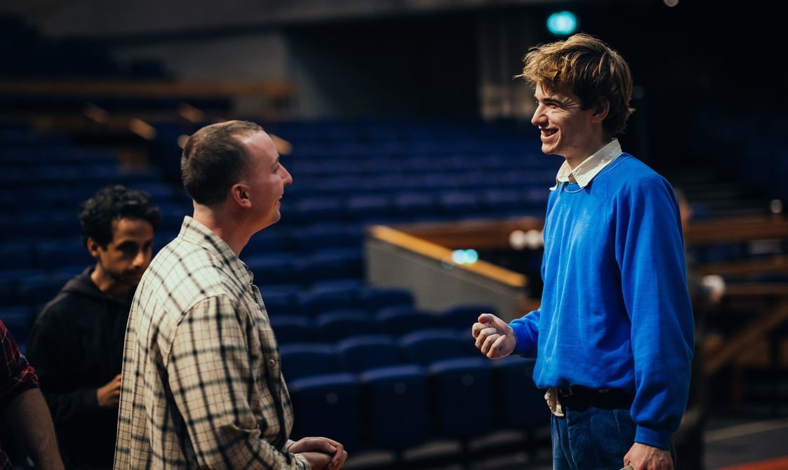 Two people talk, smiling at each other in a pleasant exchange. The person on the left wears a checkered shirt and clasps their hands together. The person on the right wears a bright blue jumper.