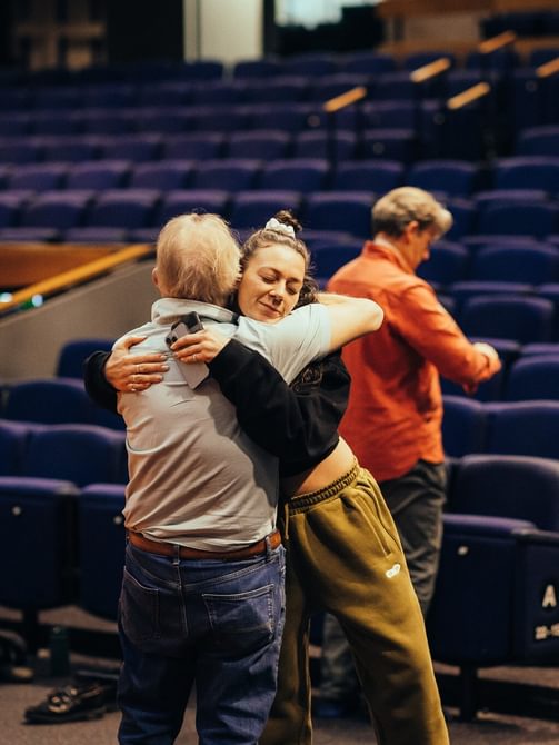 A man and woman stand together, embracing each other in a hug. They are in the auditorium, behind them are ranks of blue seats. The woman is smiling with her eyes closed.