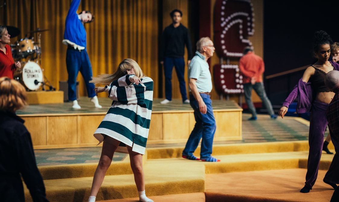 Actors warm up on the stage, they are all in motion and holding various poses. The stage is arranged in many hexagonal layers, carpeted in red, orange and yellow. The actors are wearing relaxed clothing. At the front of the image is a woman, wearing a large shirt, swaying as both her arms cross over one another.