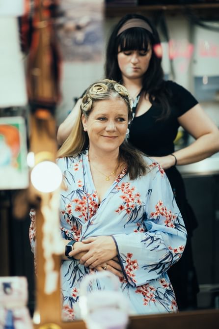 A woman getting ready in the dressing room smiles into the mirror. She is wearing a floral blue dressing gown and half of her hair is up in pin curls. Behind her another woman, dressed in black clothes, is doing the actor’s hair.