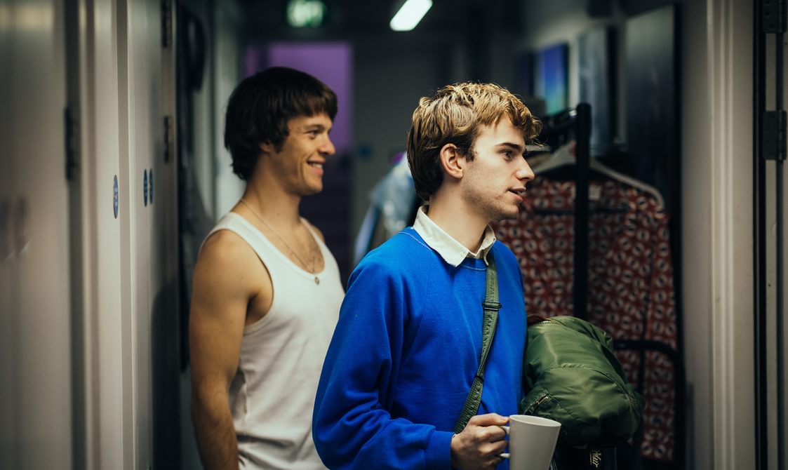Two actors backstage in a busy corridor of costume rails. The actor in the front of the image carries a white mug in one hand and his coat in the other. He is wearing a striking blue jumper. The actor behind him smiles into a nearby door. He is wearing a white tank top.