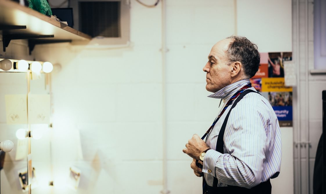 A man wearing a blue and white stripped shirt looks in the mirror as he ties his tie. He has a concentrated expression. The mirror is lit by dressing room-style light bulbs.