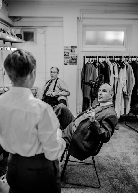 Three actors are talking casually in the dressing room. Two of the actors are seated, looking towards the third actor whose back is to the door. Behind them is a rail of costumes. They all wear smart shirts.