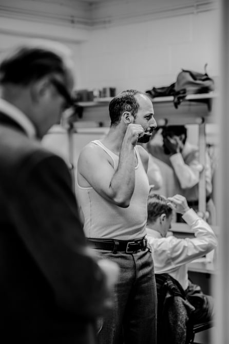 A man in a white tank top brushes his teeth in a busy dressing room. Around him, other actors are getting dressed.