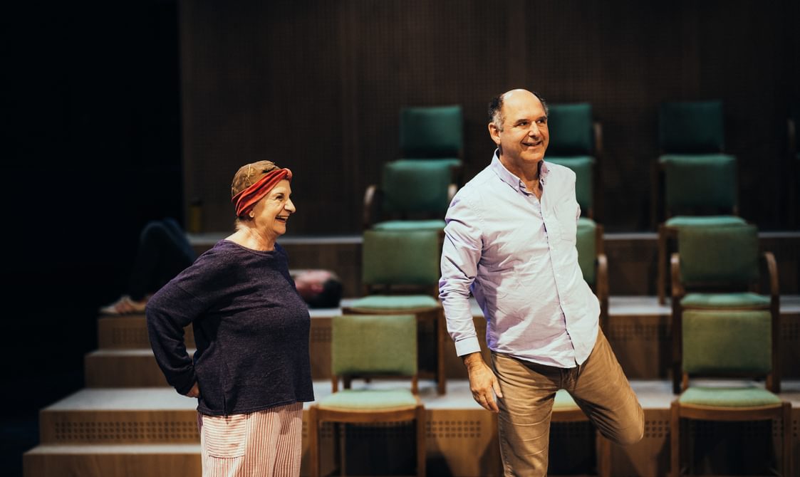 Two actors warm up on stage, smiling as they stretch. The woman on the left has her hair in pin curls and wears a navy top with white and red stripped trousers. The man on the right, who wears a blue shirt and chinos, stretches one leg behind him. In the background we can see ranks of green chairs.