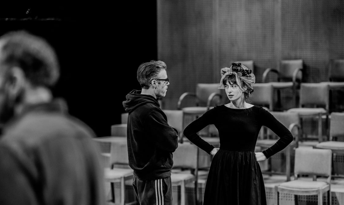 A man and a woman wearing dark rehearsal clothes stand on stage looking at each other. The man has his arms crossed and the woman has her hands on her hips. She looks at him with a suspicious expression. Behind them are ranks of chairs.