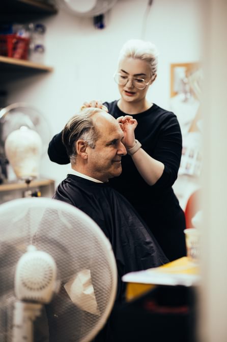 An actor sits in a dressing room while a woman adjusts his wig. He wears a hairdressing cape and smiles into the mirror in front of him. Several fans are dotted around the room.