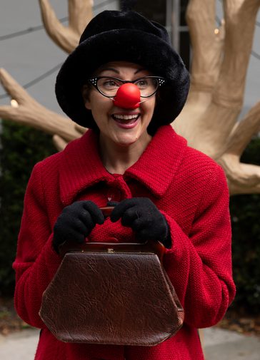 An actress is dressed as a Ladybird, with a red nose, red coat and black hat and gloves. She holds a a handbag clutched to her chest smiling pensively.