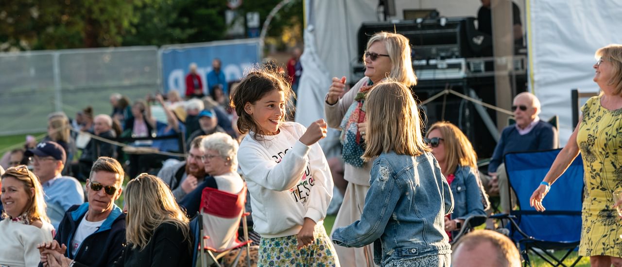 Two girls dance together on the park. They are smiling. Behind them another woman dances, and lots of other people sit on chairs looking towards a stage.