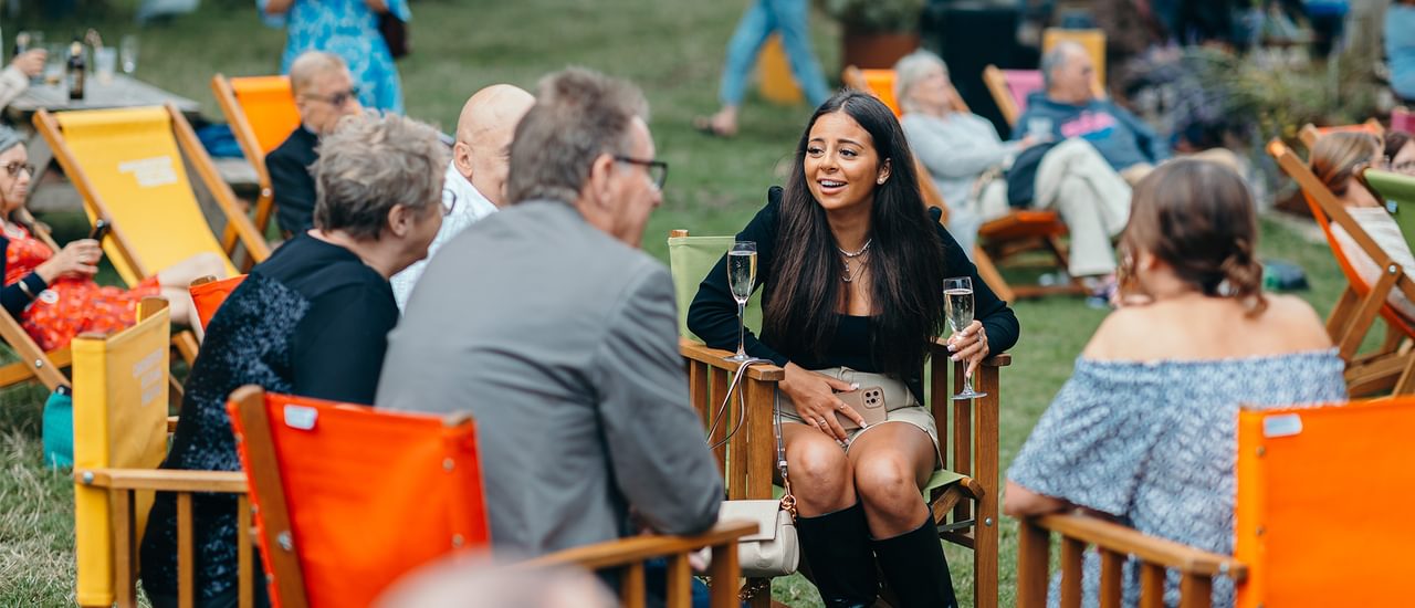 A group of people sit on colourful deckchairs in the park, chatting and smiling.