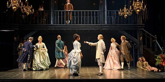 A formal dance scene. In the centre, a young actor presents their hand to a young woman in a blue dress, offer her a dance. Behind them, couples dance round in circles together. A young actor looks down on the scene from a balcony.