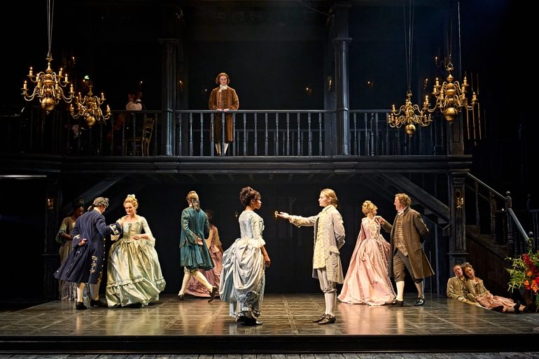 A formal dance scene. In the centre, a young actor presents their hand to a young woman in a blue dress, offer her a dance. Behind them, couples dance round in circles together. A young actor looks down on the scene from a balcony.