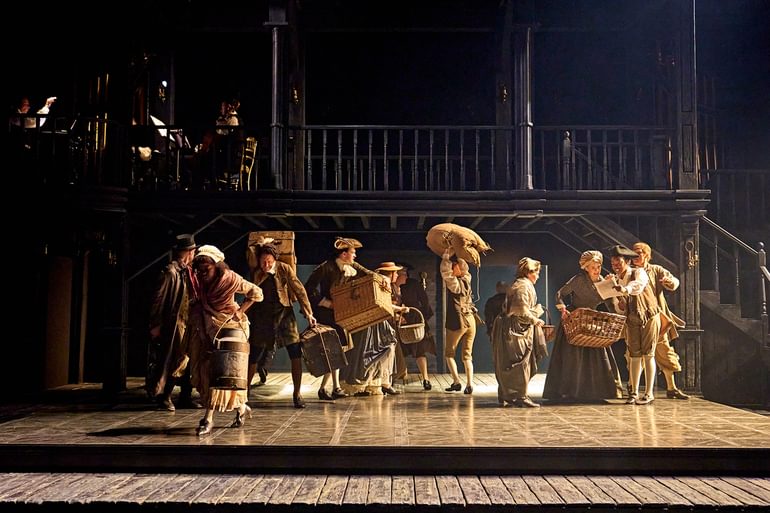 A group of actors stand on a wooden-floored stage, carrying sacks and baskets. They are moving in different directions, some holding things over their heads. On the right, a smiling young man shows a paper to a woman.