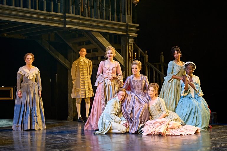 A group of people dressed in colourful dresses and jackets stand and sit together on stage. In the centre an older woman in a frilly purple dresses sits on a chair, looking proud, with two younger actors sat on the floor leaning against her dress. To the right two women in blue dresses lean against each other, hodlign hands. In the background stands a man in a golden jacket with buckles down the front. Off on her own stands a maid in a plain grey dress, with a serious expression.