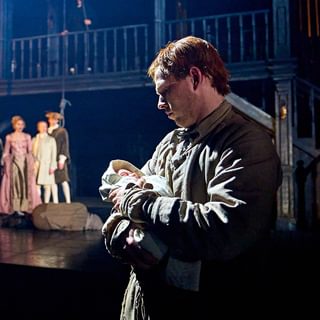 A man stands in front of the camera, holding a newborn baby who is wrapped in a blanket. The man looks down at the baby with a serious expression, and they are lit in white light. In the background, a group of people in long flowy dresses and smart jackets stand together in a freeze frame. A man has collapsed on the floor in front of them.