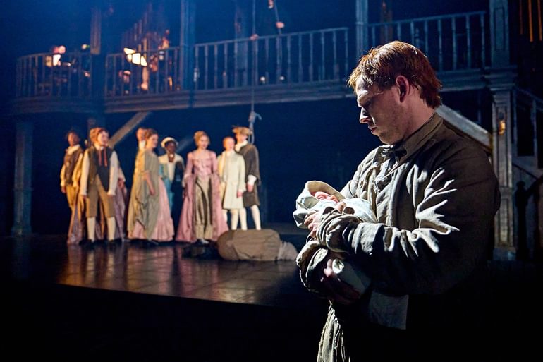 A man stands in front of the camera, holding a newborn baby who is wrapped in a blanket. The man looks down at the baby with a serious expression, and they are lit in white light. In the background, a group of people in long flowy dresses and smart jackets stand together in a freeze frame. A man has collapsed on the floor in front of them.